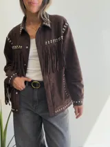 Chaqueta de efecto gamuza en color chocolate, con diseño de flecos en el pecho y mangas. Presenta detalles de tachas metálicas plateadas a lo largo de los hombros, solapas y bolsillos. Cuenta con cierre frontal de botones y forro interior.
