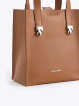 Cartera tipo tote pequeña de cuero color tostado, con doble asa superior y herrajes metálicos plateados en forma de llama en la base de las manijas. Presenta el logo de la marca estampado en la parte inferior frontal.