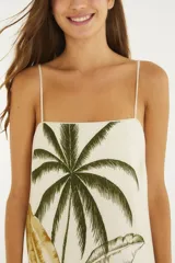 Vestido largo blanco de algodón con tirantes finos, escote recto y corte amplio en línea A. Presenta un estampado posicional tropical con palmeras, hojas de banano y arquitectura colonial.