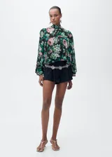 Blusa de gasa con estampado floral en tonos rosa, verde y negro. Tiene cuello alto, mangas largas con volantes y cierre de lazo en la parte posterior.