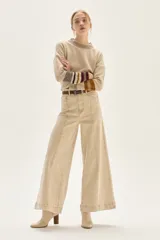 Pantalón de jean color beige claro, tiro alto, de corte ancho (wide leg) y largo hasta el tobillo con dobladillo. Presenta costuras verticales marcadas en el frente de las piernas.