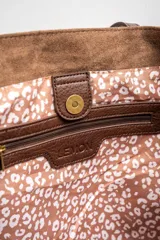 Bolso tipo tote de gran tamaño confeccionado en gamuza sintética color tostado. Presenta un diseño minimalista con asas largas de hombro y una pequeña placa metálica frontal con el logo de la marca.