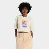 Remera cropped de algodón color crema con mangas cortas y cuello redondo. Presenta un estampado gráfico central que muestra un par de zapatillas deportivas sobre un fondo de bloques de color degradado en tonos amarillo, rosa, morado y azul.