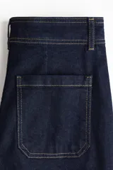 Jean de tiro alto con corte acampanado desde la rodilla. Confeccionado en denim azul oscuro con costuras a contratono, presenta bolsillos tipo parche en la parte frontal y cierre con botón metálico.