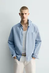 Camisa de hombre de manga larga, corte relajado (relaxed fit), color celeste con patrón de cuadros pequeños en azul y rojo. Presenta cuello abotonado (button-down), bolsillo de parche en el pecho y cierre frontal de botones.