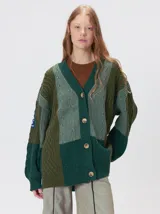 Cardigan de punto grueso con diseño patchwork en tonos verdes, cuello en V y cierre frontal con botones. Presenta mangas largas abullonadas, corte oversize y parches decorativos en el pecho y hombro.