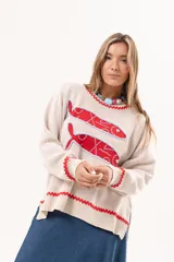 Sweater tejido color crudo con cuello redondo y mangas largas. Presenta dos peces rojos con detalles en azul bordados en el frente. Tiene ribete ondulado rojo en cuello, puños y ruedo.