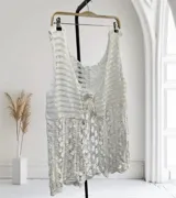 Chaleco largo sin mangas, color blanco, tejido a crochet con un patrón calado que incluye motivos florales y cierre frontal anudado.