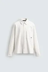 Camisa de corte relajado confeccionada en tejido de algodón, con diseño de rayas verticales. Presenta cuello solapa, cierre frontal de botones, manga larga con puños abotonados y un bolsillo plastrón en el pecho con un pequeño bordado decorativo.