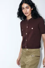 Chaqueta de punto color marrón oscuro, con cuello redondo, manga corta y cierre frontal de botones dorados. Presenta dos bolsillos falsos con botón dorado en la parte delantera superior.