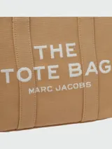 Bolso tipo tote bag de Marc Jacobs en color beige, confeccionado en lona resistente. Presenta el texto "THE TOTE BAG" y "MARC JACOBS" impreso en la parte frontal en color negro. Cuenta con asas superiores y una correa de hombro ajustable y extraíble, además de ribetes laterales negros con inscripciones.