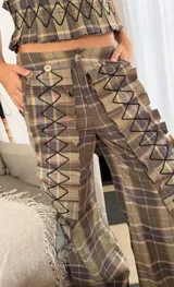Pantalón de corte amplio con estampado de cuadros escoceses en tonos azul y verde. Presenta apliques de tela plisada con costuras decorativas en zigzag a lo largo de las piernas, tajos laterales y bolsillos funcionales.