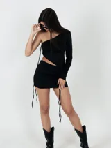 Conjunto de dos piezas color negro, compuesto por un top asimétrico de un solo hombro con manga larga y una minifalda ajustada. Ambas prendas cuentan con detalles de tiras laterales ajustables.