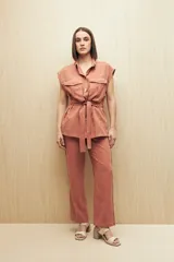 Chaleco color beige con cuello camisero, sin mangas, cierre frontal con botones y cinturón de lazo. Presenta bolsillos de parche en el pecho y pinzas verticales en el frente.