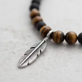 Collar largo compuesto por cuentas de piedra natural ónix negro y ojo de tigre, con dije central de pluma en acero y argolla metálica.