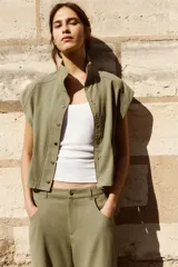 Chaleco de cuello alto y manga sisa, color verde militar, con elásticos fruncidos en la cintura y cierre frontal de botones metálicos a presión.