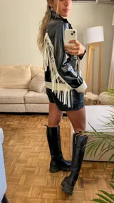 Campera negra de cuero sintético con cierre frontal, detalles en blanco en cuello, puños y ruedo, y flecos blancos en las mangas.