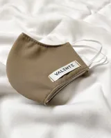 Tapabocas de tela color marrón claro con elástico blanco en los laterales para sujetar a las orejas y etiqueta con la marca Valente.