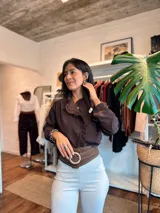 Camisa de color marrón oscuro con cuello mao y detalle de volados a lo largo de la abertura frontal. Presenta mangas largas con puños rectos y cierre frontal mediante botones ocultos.