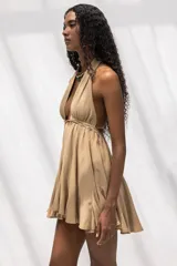 Vestido corto color beige con escote halter profundo, espalda descubierta y falda con vuelo. Presenta un diseño fluido y ligero con detalle de frunces en la cintura.