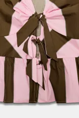 Camisa de rayas verticales rosadas y marrones, con escote en V, mangas corta abullonadas y cierre frontal con múltiples lazadas del mismo tejido. Presenta bajo con acabado de volante.