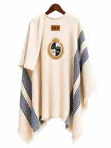 Ruana de tejido de punto en color beige con diseño de rayas verticales azules en los laterales y flecos en el ruedo. Presenta un escudo bordado central con motivos dorados y azules, y una etiqueta de cuero con la marca Bagual en la parte superior.