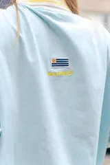 Polo de manga larga en color marrón con rayas horizontales azules en las mangas. Presenta cuello tipo polo en azul con detalles de rayas, cierre frontal parcial con botones y estampado frontal con la frase 'From Uruguay to the World'. Incluye un parche bordado en el pecho.