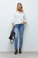 Blusa de color crudo con escote en V y mangas tres cuartos abullonadas. Presenta un delicado diseño de flores bordadas en tonos azules y amarillos distribuidas por toda la prenda.