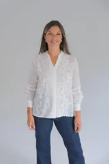 Blusa blanca de manga larga con cuello mao y cierre frontal de botones ocultos. Presenta un delicado diseño con bordados florales y detalles calados verticales en el frente.