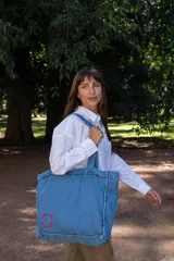 Bolso tipo tote confeccionado en tela denim de color azul claro. Presenta un diseño amplio con costuras visibles, doble asa de hombro y un detalle bordado circular en color rojo en la parte inferior.