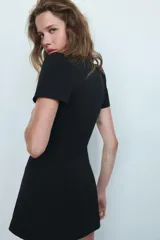 Vestido corto de color negro con escote en pico profundo, mangas cortas abullonadas y detalle de lazo frontal. Presenta un diseño entallado con cierre de cremallera oculto en la espalda.