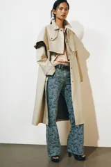 Gabardina larga de corte recto y diseño minimalista en color beige, con cuello solapa, manga larga y cierre frontal con botones. Cuenta con una capa desmontable en el mismo tejido y una solapa decorativa en el pecho.