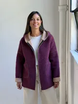 Tapado de paño color magenta con capucha integrada, cierre frontal y bolsillos laterales. Presenta detalles en color beige en el interior de la capucha, puños y bordes de los bolsillos.