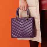 Cartera de mano color púrpura, con diseño acolchado en símil cuero soft y costuras a tono. Tiene acceso con cierre y broches imantados, herrajes dorados y correa regulable para usar como bandolera. El interior está forrado en tela de algodón y poliéster, con un bolsillo con cierre y dos abiertos.