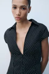 Camisa entallada de popelín negro con estampado de lunares blancos, escote en pico, cuello solapa y mangas muy cortas. Cuenta con costuras marcadas y cierre lateral con cremallera oculta.