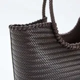 Bolso de mano grande con diseño rectangular y textura trenzada en color marrón oscuro. Cuenta con dos asas circulares trenzadas integradas en la estructura.