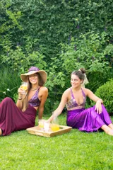 Vestido largo con estampado tie-dye en tonos violeta y fucsia, escote en V con detalles bordados dorados, cut-outs laterales bajo el busto y tirantes finos que se cruzan y atan en la espalda.