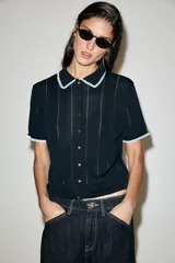 Chaqueta de punto de manga corta con cuello polo y cierre frontal de botones, en color negro con detalles de puntilla blanca en el cuello y los puños.