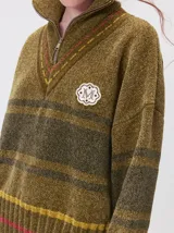Pullover de textura tipo terciopelo con cuello alto y medio cierre frontal. Presenta diseño a rayas en tonos tierra con detalles de pespuntes en el cuello y parche bordado en el pecho. Posee puños y ruedo acanalados con corte oversize.