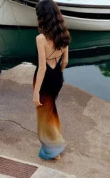 Vestido largo de seda o material similar, con tirantes finos y escote drapeado tipo cowl neck. Presenta un efecto degradado de color que va del negro/marrón oscuro en el busto, pasando por tonos naranjas y amarillos, hasta terminar en un azul vibrante en el ruedo.