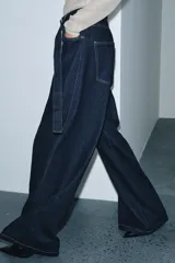 Jeans de tiro alto con pierna ancha, confeccionados en denim azul oscuro. Presentan cinco bolsillos, trabillas en la cintura y un cinturón ajustable del mismo tejido con hebilla metálica. Incluyen pespuntes marcados a tono y cierre frontal con cremallera y botón.