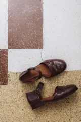 Zapato de cuero color borgoña, con puntera redondeada y diseño trenzado. Presenta una tira sobre el empeine con hebilla metálica negra.