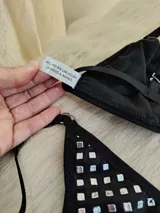 Conjunto de bikini negro con apliques de tachas cuadradas plateadas. El corpiño es de estilo triangular con una argolla central metálica y breteles regulables. La bombacha es de corte colaless con tiras laterales ajustables y argollas metálicas en los extremos.