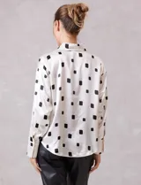 Camisa de satén con estampado de rectángulos negros sobre fondo blanco. Presenta cuello clásico con solapa, cierre frontal con botones metálicos y mangas largas con puños rectos.