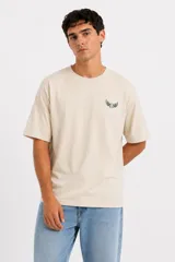 Remera de algodón color beige, de manga corta, cuello redondo y corte holgado. Presenta un pequeño estampado verde en el pecho con diseño de pelota con alas.