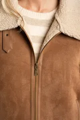 Chaleco estilo gamulán de corte oversize, confeccionado en material tipo gamuza color camel con interior y bordes de corderito sintético color crema. Presenta cierre frontal completo, bolsillos laterales y detalles de tiras decorativas en los hombros.