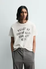Camiseta de manga corta y cuello redondo, color blanco roto con efecto lavado y desgastado. Presenta un estampado frontal en negro con texto de estilo universitario que incluye "PRINCE ST.", "GROOVE ALLEY", "SOHO, NYC" y el número "95".