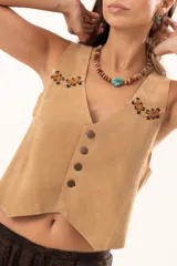 Chaleco de gamuza color beige con estilo western, presenta cierre frontal con botones y detalles de ojalillos metálicos con cordones de cuero entrelazados en la parte superior del frente.
