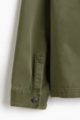 Sobrecamisa de lona de algodón en color verde militar, con cuello clásico, cierre frontal de botones y tres bolsillos de parche frontales. Presenta mangas largas con puños abotonados y un diseño de corte recto.
