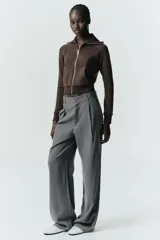 Pantalón gris de tiro alto con bolsillos delanteros y traseros, detalle de pliegues frontales y cierre frontal con cremallera y botón.